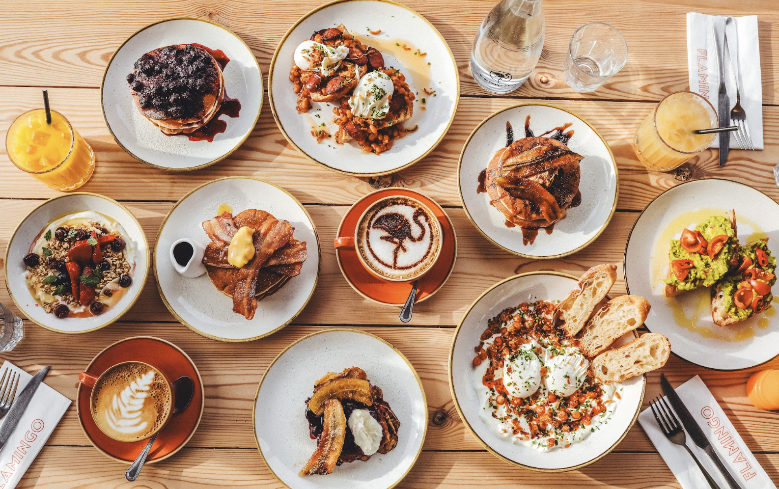 A variety of brunch dishes from the Flamingo By Day Menu are arranged on a wooden table. Plates include pancakes, avocado toast, eggs, fruit, and pastries. Two glasses of orange juice, cups of coffee, and silverware are also visible, along with branded napkins.
